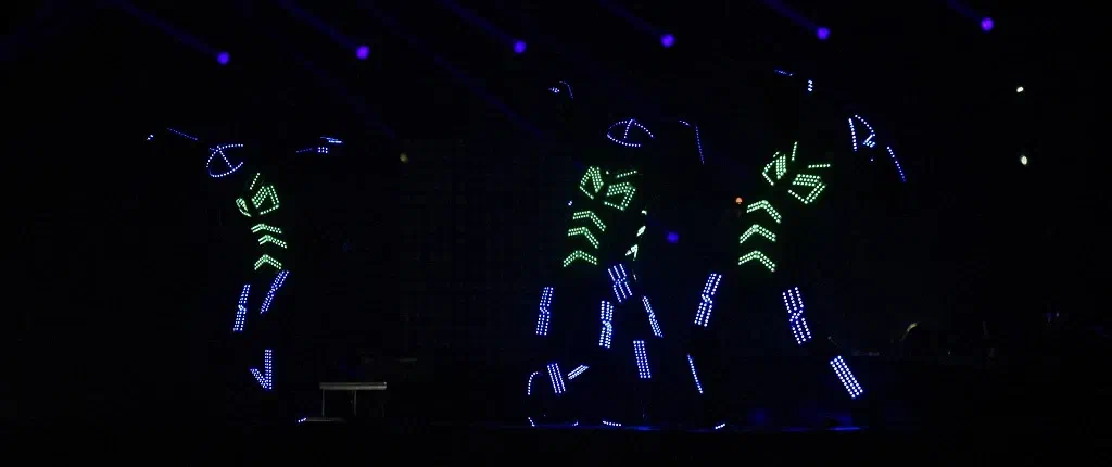 Three Tron dancers in LED suits with glowing green chevrons and blue-lit arms and legs, dancing on a dark stage under purple spotlights