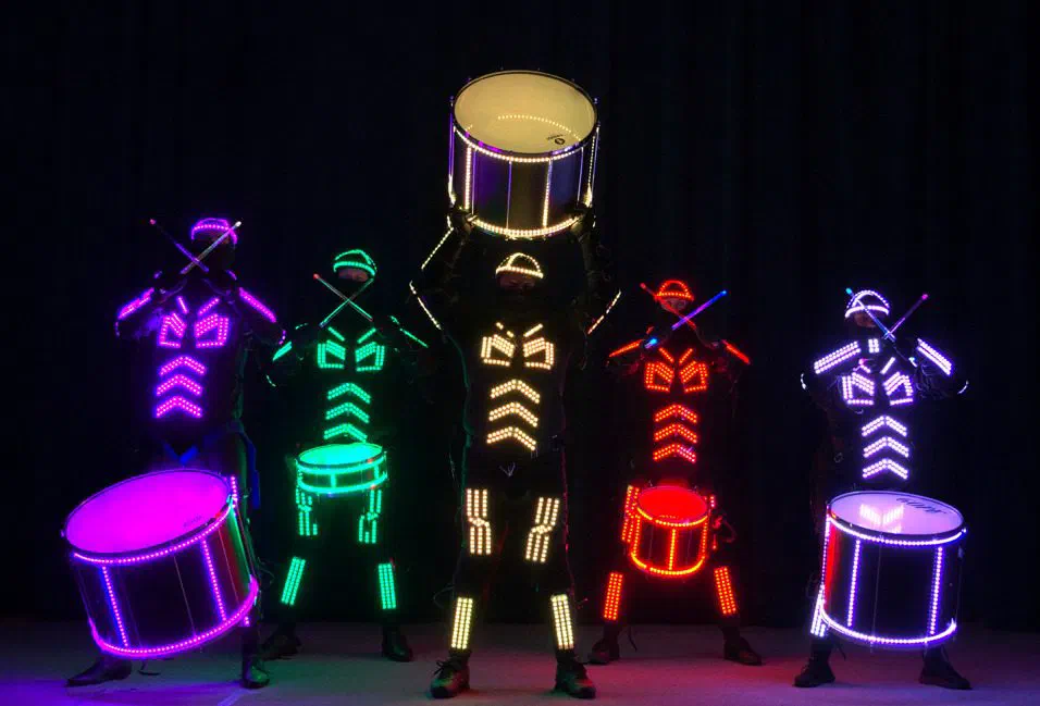 Päť bubeníkov Tron Drummers v žiariacich LED kostýmoch so svietiacimi bubnami, jeden bubon zdvihnutý nad hlavou
