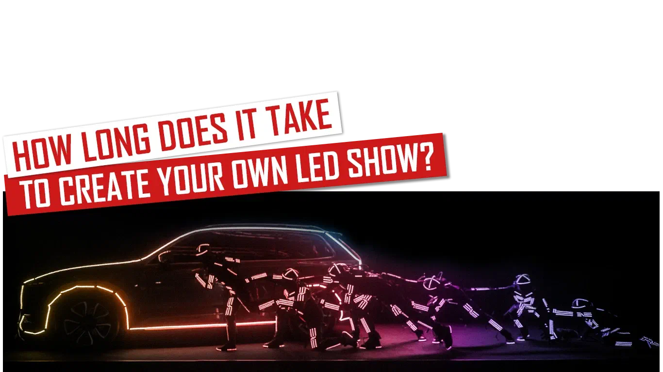 How long does it take to create your own LED show? – red and white banners above a row of dancers in glowing pink and purple LED suits leaning away from a car outlined in LED light, as if pulling it, in the dark