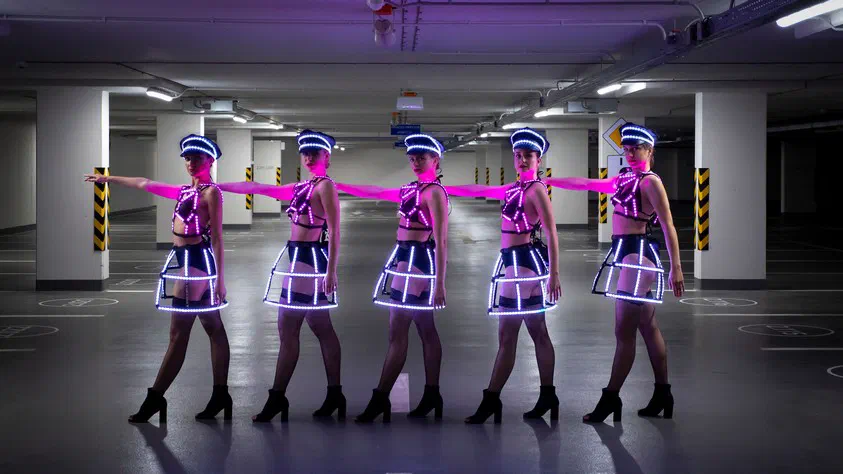Five Tron Girls dancers in the black version of the Dolls costumes – LED caps, harnesses and cage skirts glowing white and pink – in a row in an underground car park, their arms stretched out