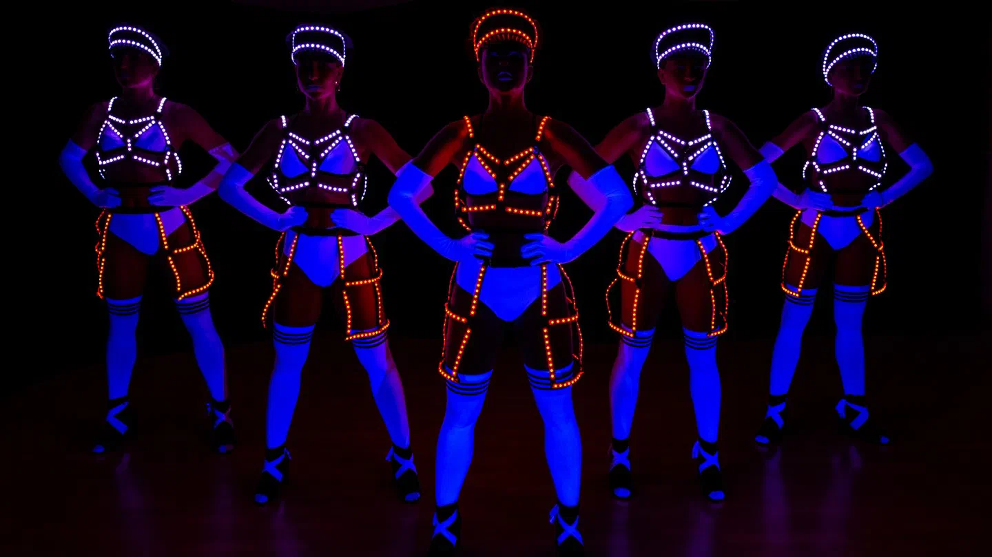 Five Tron Girls dancers in white Dolls costumes glowing blue under UV light, with white and orange LED harnesses, caps and cage skirts, standing with hands on hips in the dark