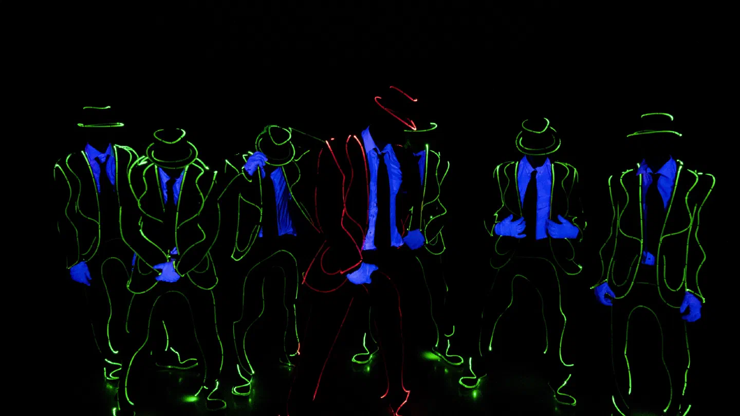 Tron Man dancers in suits and hats outlined by glowing green lines, with blue shirts and gloves under UV light, one in the middle outlined in red, in the dark