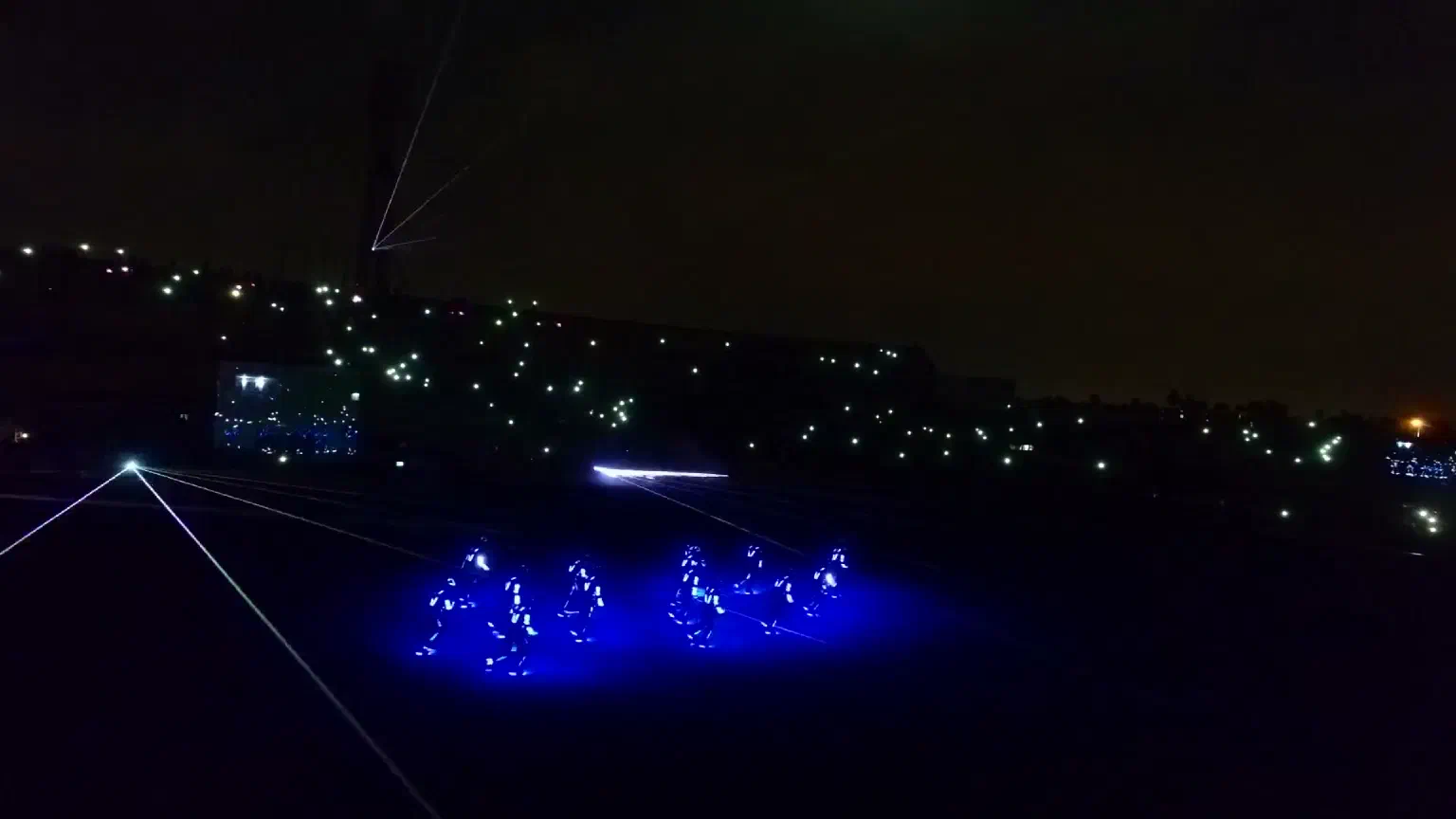 Tron Dance show: dancers in blue LED suits on a stadium field at night, with white laser beams and lights in the stands