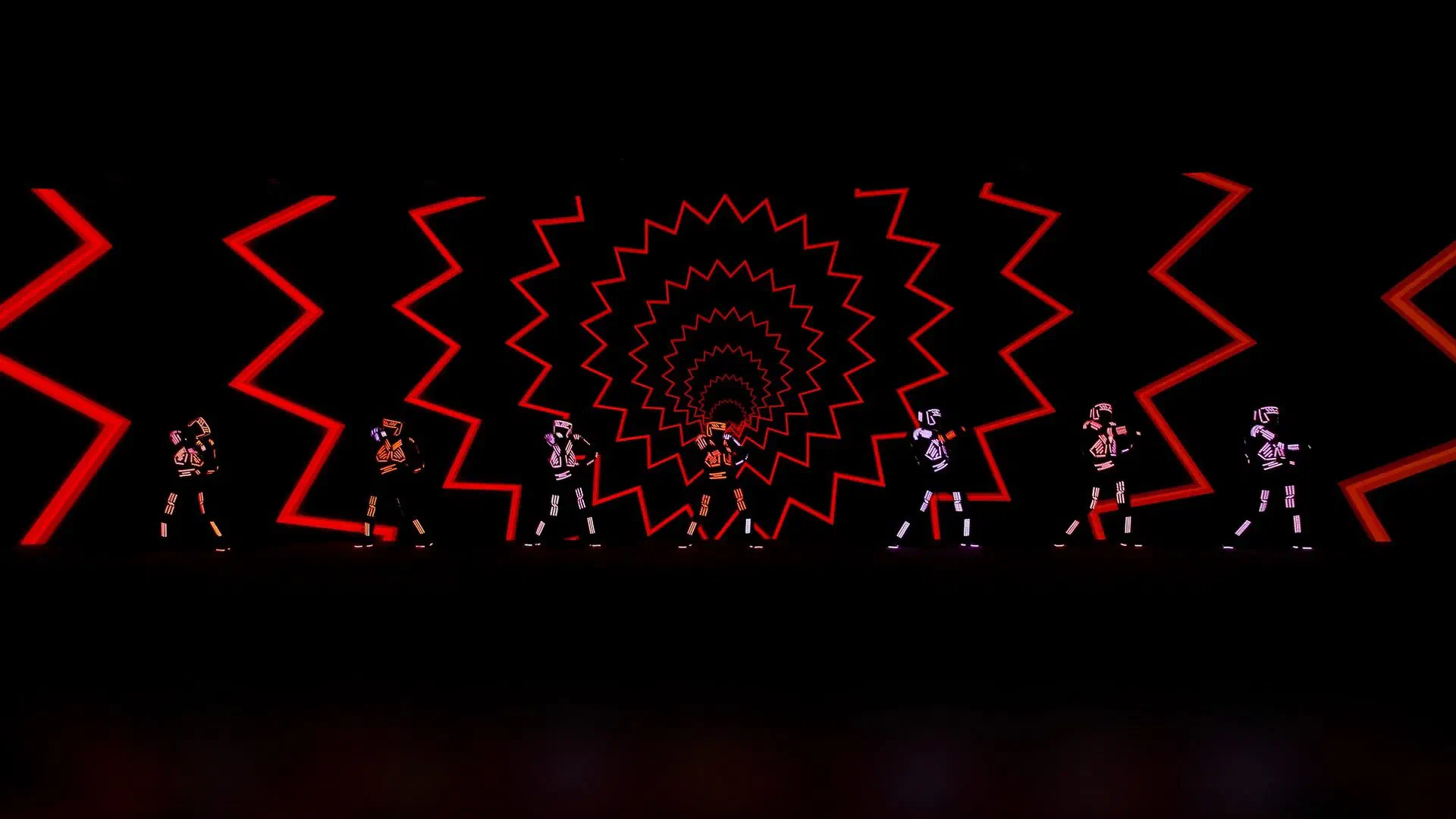 Seven Tron Dance Gen II dancers in glowing LED suits in front of a red zigzag LED-wall graphic