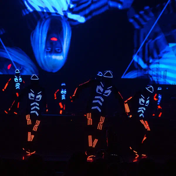 Tron Dance: five dancers in blue and orange LED suits on stage before a video screen, audience in the foreground