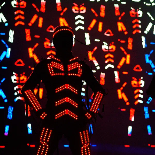 Tron Dance: dancer in an orange-red LED suit, hands on hips, before a wall of projected multicoloured LED figures