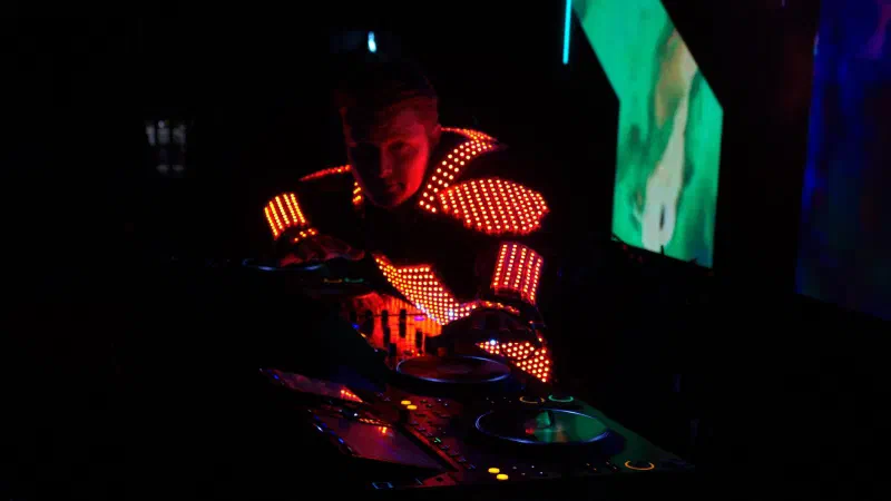 Tron DJ in an orange-red LED pixel suit mixing at the decks, with visuals on LED screens behind him