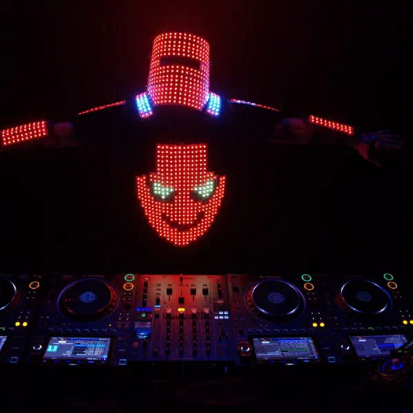 Tron DJ: DJ in a red LED helmet and smiley-face LED shirt spreading his arms behind a four-deck DJ console