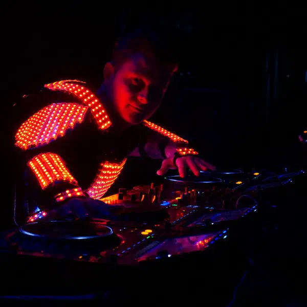 Tron DJ: DJ in an orange LED jacket mixing on the decks in a dark room, seen from the side