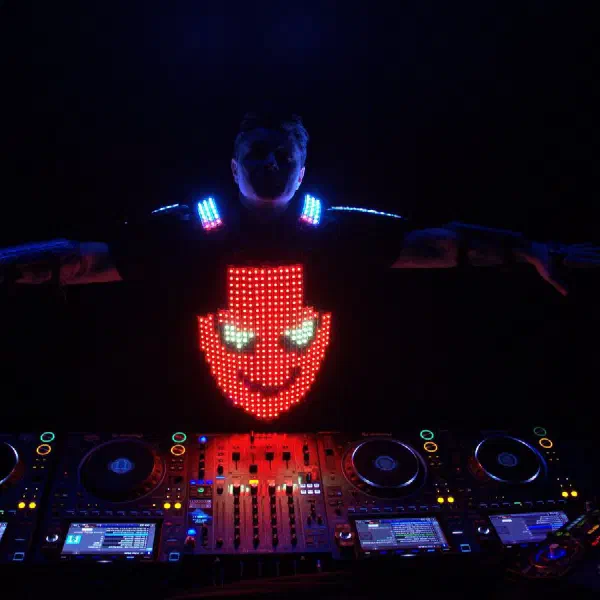 Tron DJ: unmasked DJ with arms outstretched, red LED smiley face on his chest, behind the DJ console