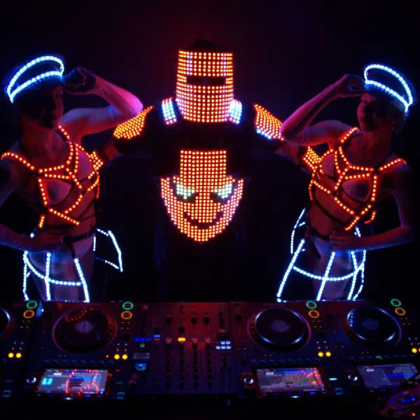 Tron DJ: helmeted LED DJ posing between two female dancers in orange LED harnesses behind the decks