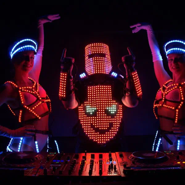 Tron DJ: helmeted DJ raising his hands as two LED dancers lift their arms beside him in an LED DJ show