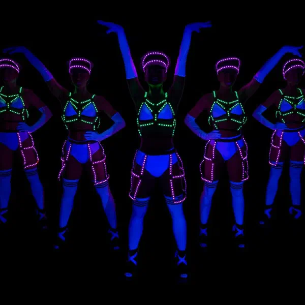 Tron Dolls: päť LED tanečníc v zelených a ružových LED postrojoch a čiapkach žiari v UV svetle na čiernom pódiu