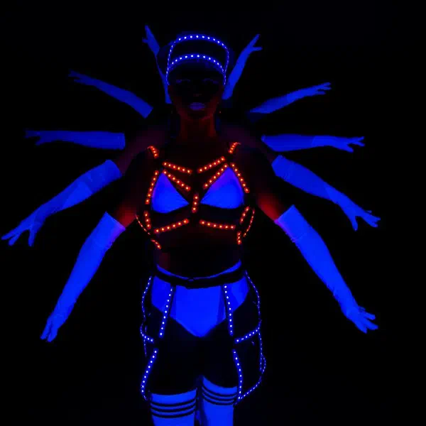 Tron Dolls: LED tanečnica v oranžovom LED postroji s mnohými rukami rozprestretými za ňou v modrom UV svetle