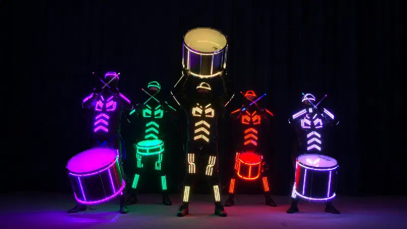 Päť bubeníkov Tron Drummers vo fialových, zelených, zlatých, červených a bielych LED kostýmoch s farebne zladenými svietiacimi bubnami, prostredný bubeník dvíha bubon nad hlavu