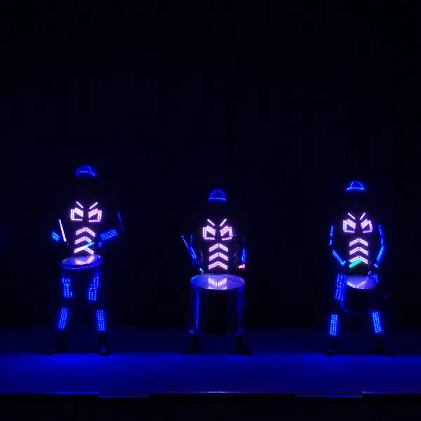 Tron Drummers: päť LED bubeníkov v modrých svietiacich kostýmoch hrá na bielo osvetlené bubny na tmavom pódiu
