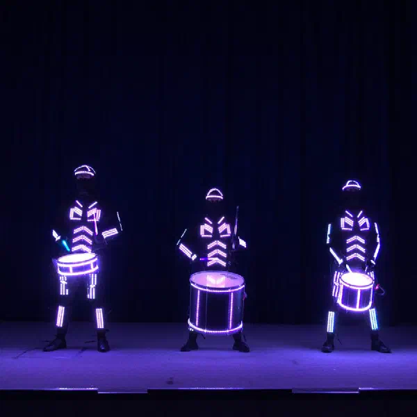 Tron Drummers: päť LED bubeníkov vo fialových a bielych svietiacich kostýmoch hrá na svietiace bubny pred tmavou oponou