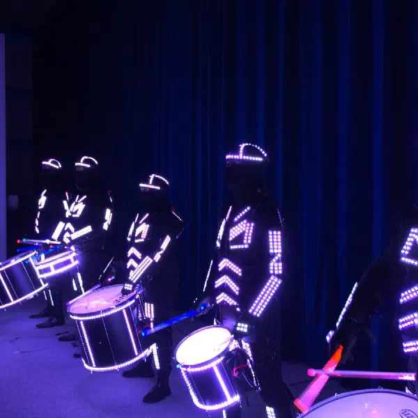 Tron Drummers: bočný pohľad na päť LED bubeníkov vo fialových LED kostýmoch a čiapkach v rade vedľa modrej opony