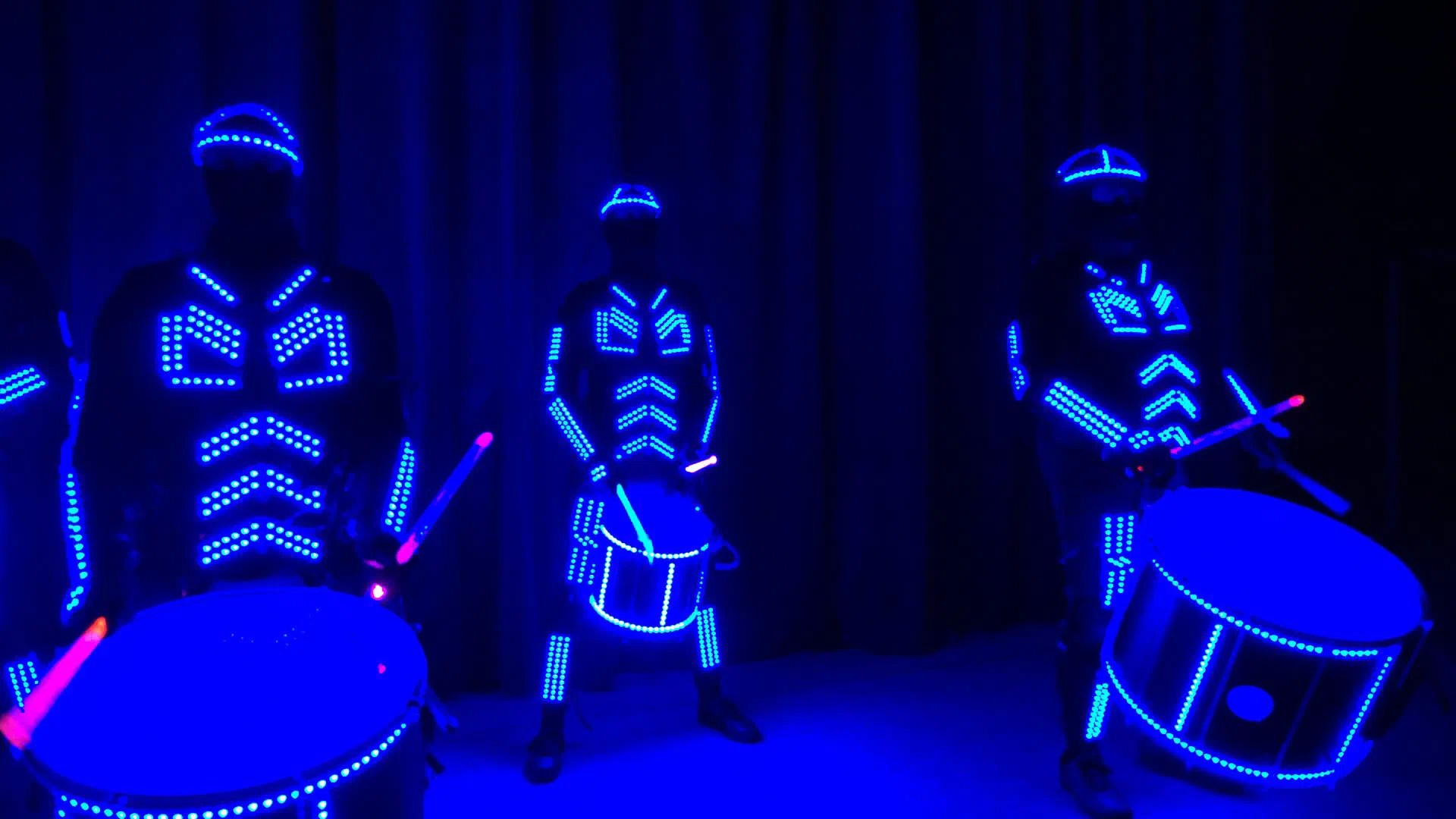 Tron Drummers v modrých LED kostýmoch so svietiacimi bubnami a paličkami pred tmavou oponou