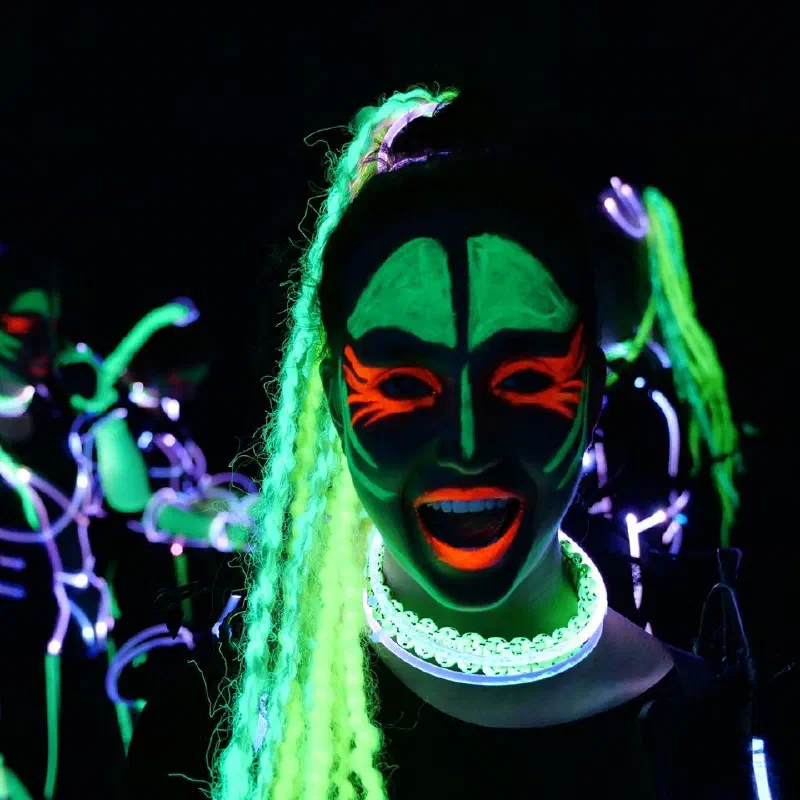 Detail tanečnice Tron Girls s neónovo zeleným a oranžovým UV make-upom, dlhými zelenými vrkočmi a svietiacim golierom, za ňou tanečnice v LED kostýmoch