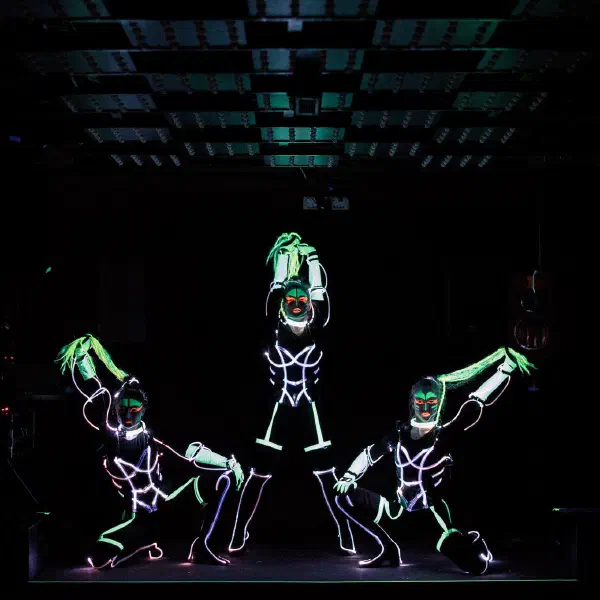 Tron Girls: tri LED tanečnice v bielych kostýmoch s neónovými drôtmi, s UV make-upom a zelenými vrkočmi na tmavom pódiu