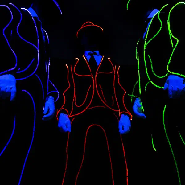 Tron Man: dancer in a red light-wire suit, hat and bow tie standing between blue and green outlined dancers