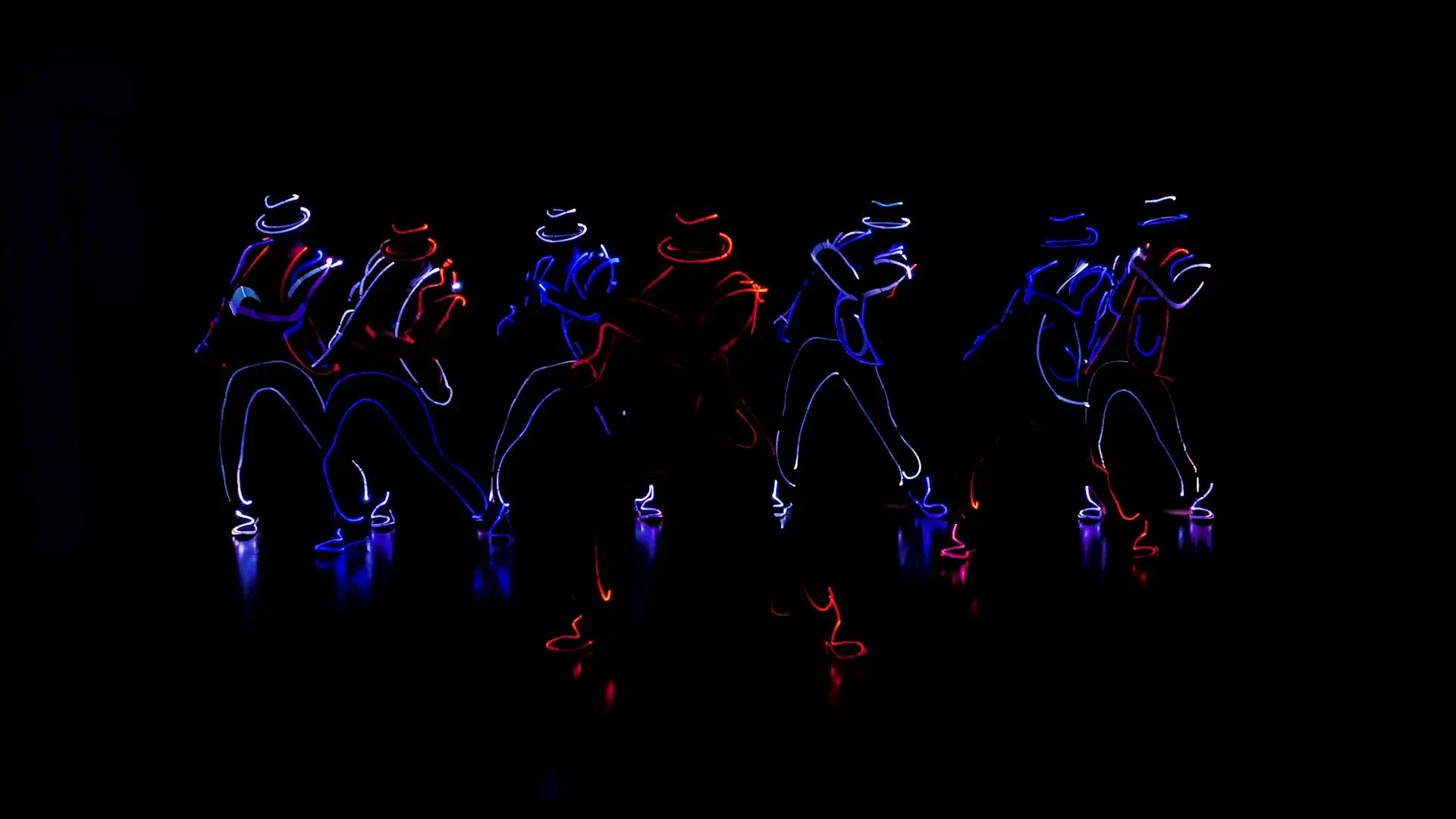 Tron Man dancers in hats and suits outlined with blue, white and red LED light, dancing in a line on a dark stage