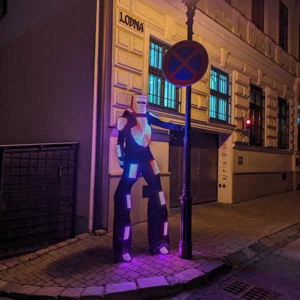 Tron Pixel Robot on stilts glowing in violet and blue pixels, leaning on a street lamp on a cobbled street corner at night