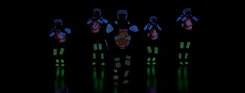 Päť LED tanečníkov Tron Pixel Show v modro-zelených pixelových kostýmoch s červeno-bielou pixelovou grafikou na hrudi na tmavom pódiu