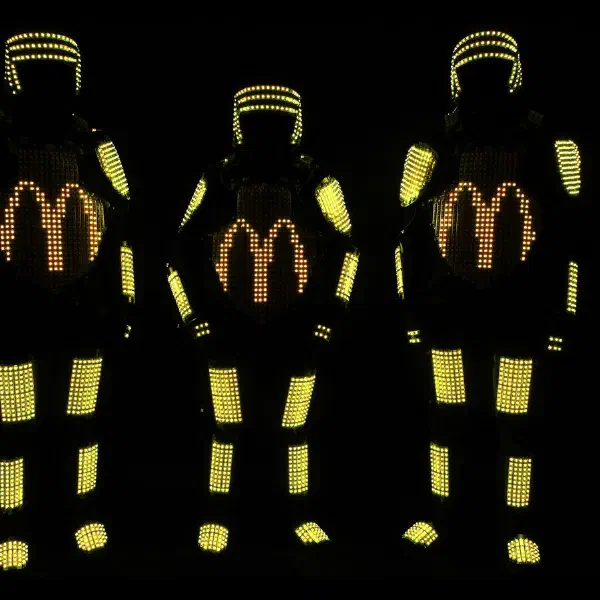 Tron Pixel Show: traja pixeloví LED tanečníci v žltých pixelových kostýmoch so zlatými oblúkmi loga McDonald's