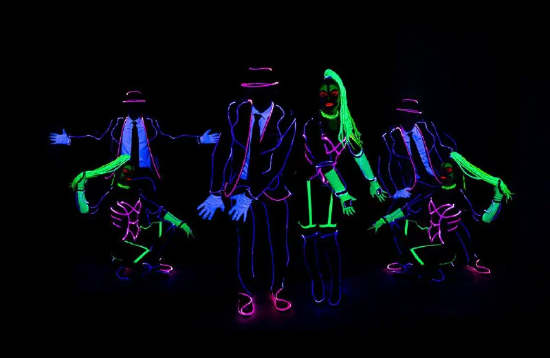 Wedding Show dancers in glowing LED outfits – men with hats and ties, women with neon green braids – posing together on a dark stage