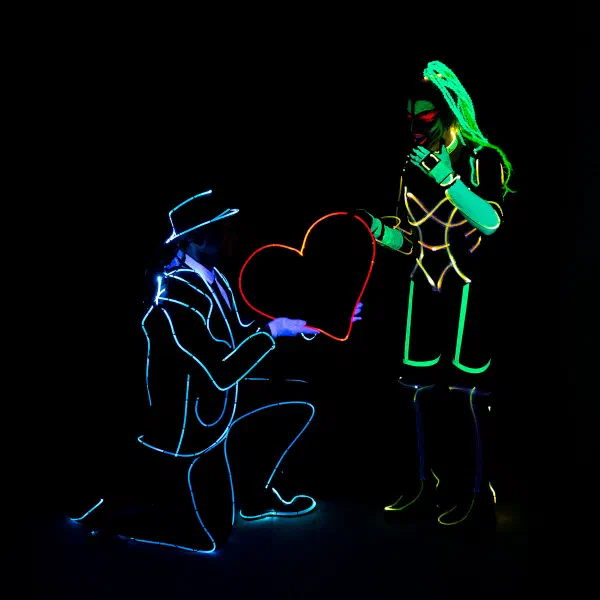 Wedding Show: man in a blue neon suit kneeling to offer a glowing red heart to a woman in green and gold light